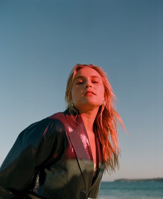 Jack river stands before a beautiful backdrop of ocean and clear blue sky. Her long blonde hair is flowing and her face is drenched in a pink light. Her holographic blazer glistens pink in the light.